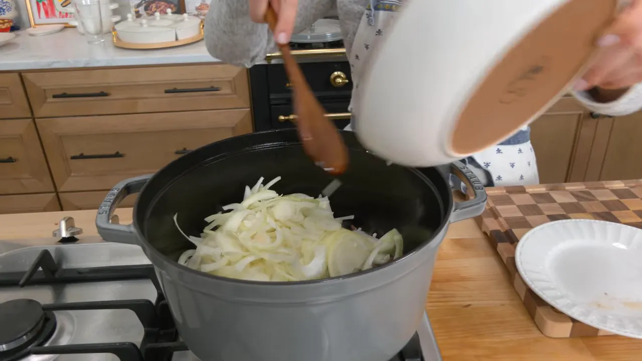 Sliced yellow onions being poured from a bowl into a large Dutch oven on the stove.
