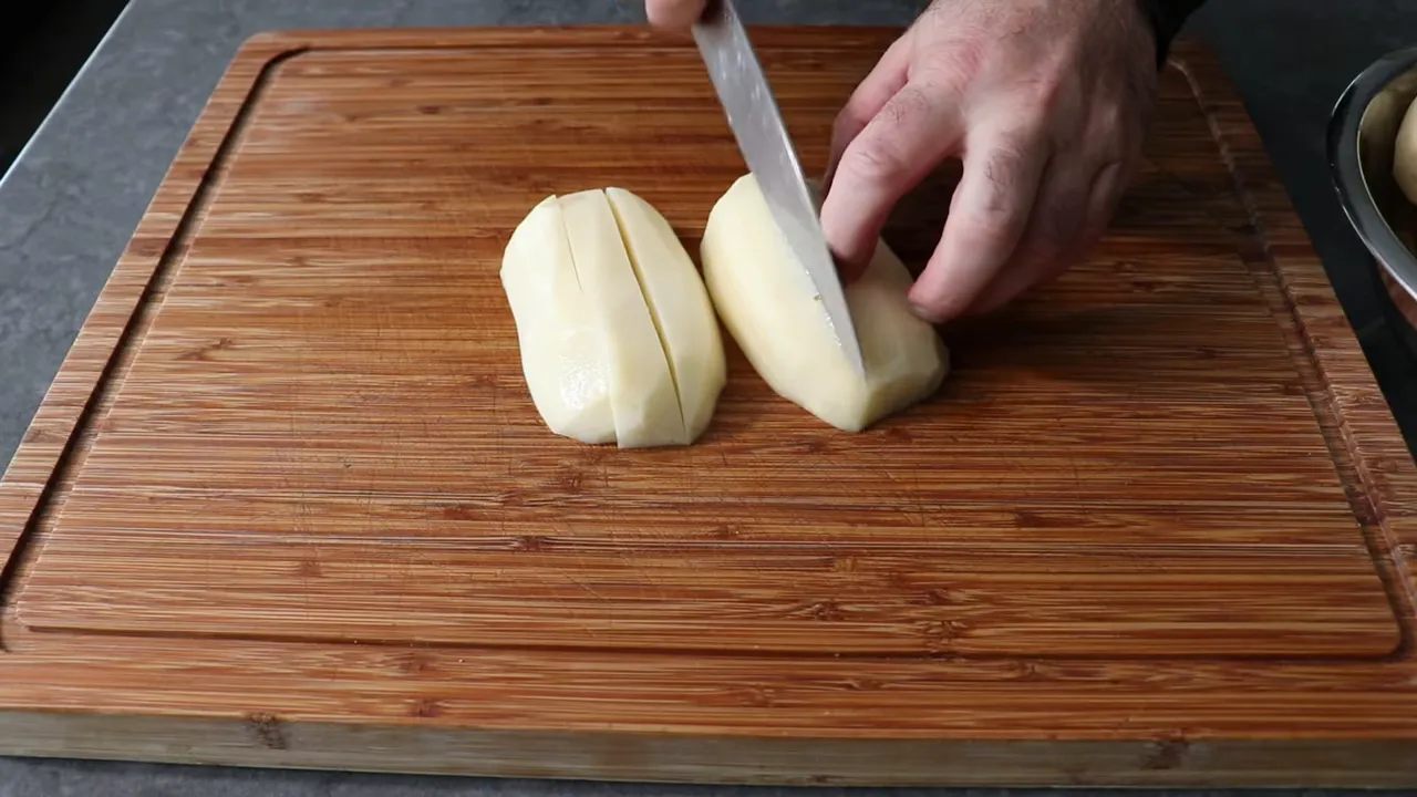 Hands cutting peeled potatoes into even thick slices on a wooden cutting board