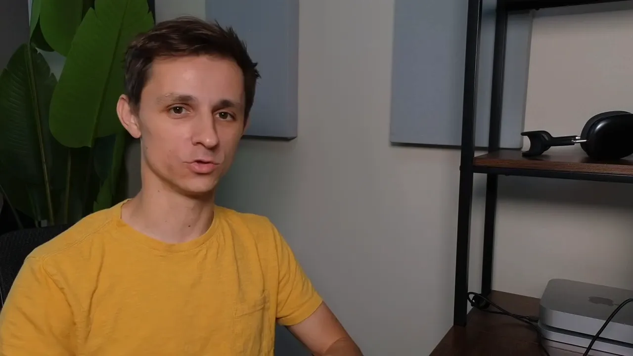 Reviewer in a yellow shirt facing the camera, speaking calmly in a home studio with shelving and headphones visible