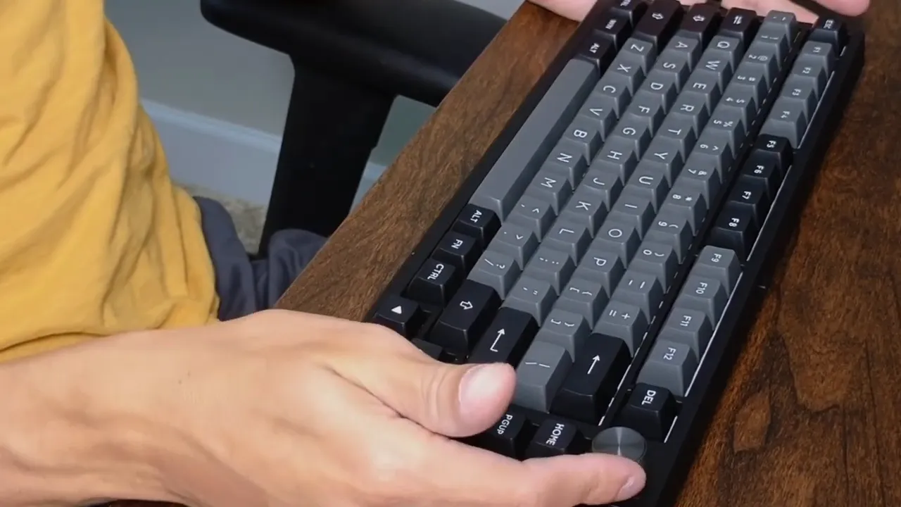 Top-right view of an Epomaker TH80 SE 75% mechanical keyboard on a wood desk showing gray PBT keycaps, arrow cluster and metal volume knob with a hand nearby.