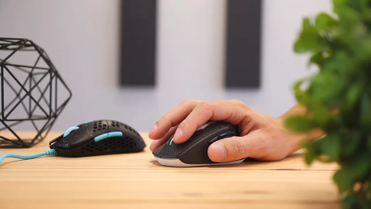 A hand resting on a Logitech mouse beside the HK Gaming Mira M for size comparison on a desk