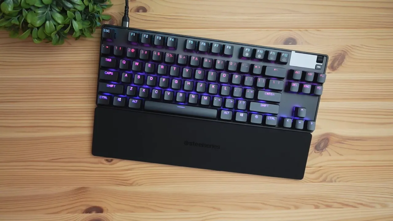 Top-down view of SteelSeries Apex Pro TKL keyboard with magnetic wrist rest showing purple and blue RGB lighting on a wooden desk.