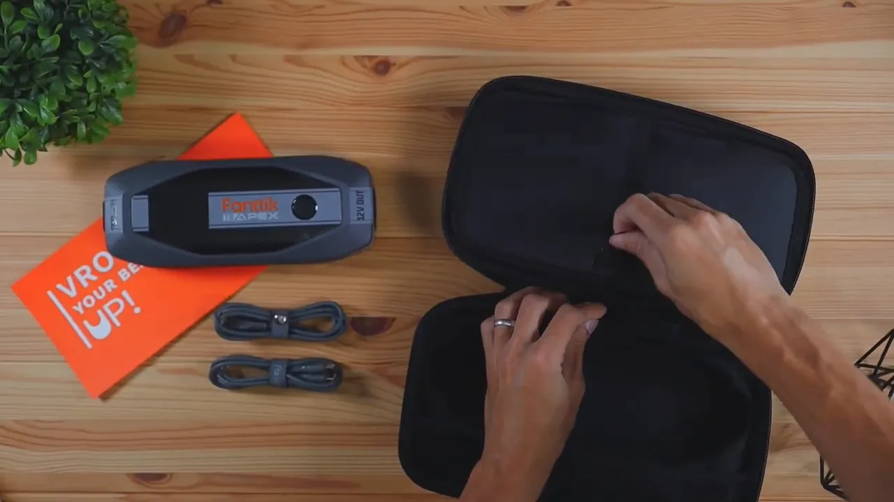 Contents of the FANTTIK T8 APEX kit arranged on a wooden table: the jump starter, two short cables, manual, and open hard shell case.