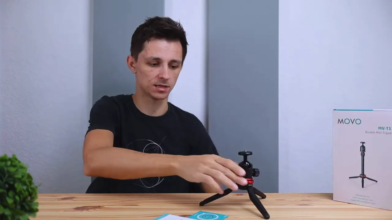 Movo MVT1 mini tripod assembled on a wooden desk as the presenter adjusts it, with the product box in the background.