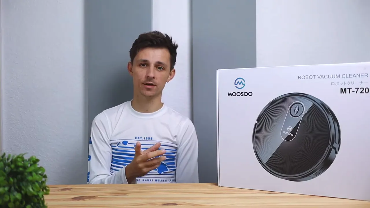 Presenter gesturing while sitting beside a boxed MooSoo MT-720 robot vacuum on a table