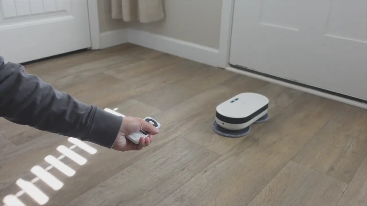 EVERYBOT Edge2 robot mop on a hardwood floor with a hand holding the remote control