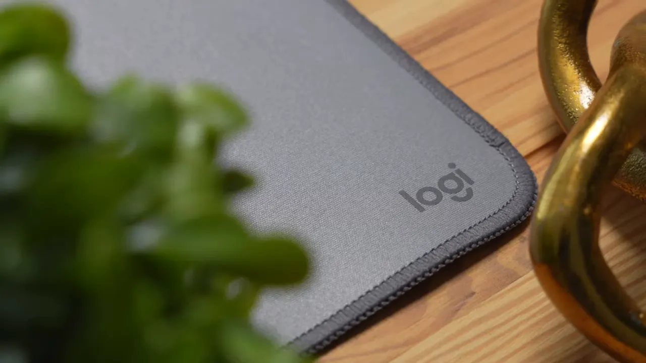Logitech desk mat corner on wood tabletop showing stitched edge, smooth gray surface, and the logi logo