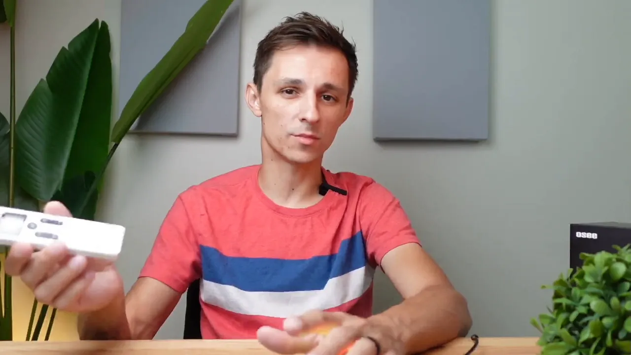 Presenter centered at a desk holding a white remote-style controller in his left hand with the OSEE box and a plant on the right.