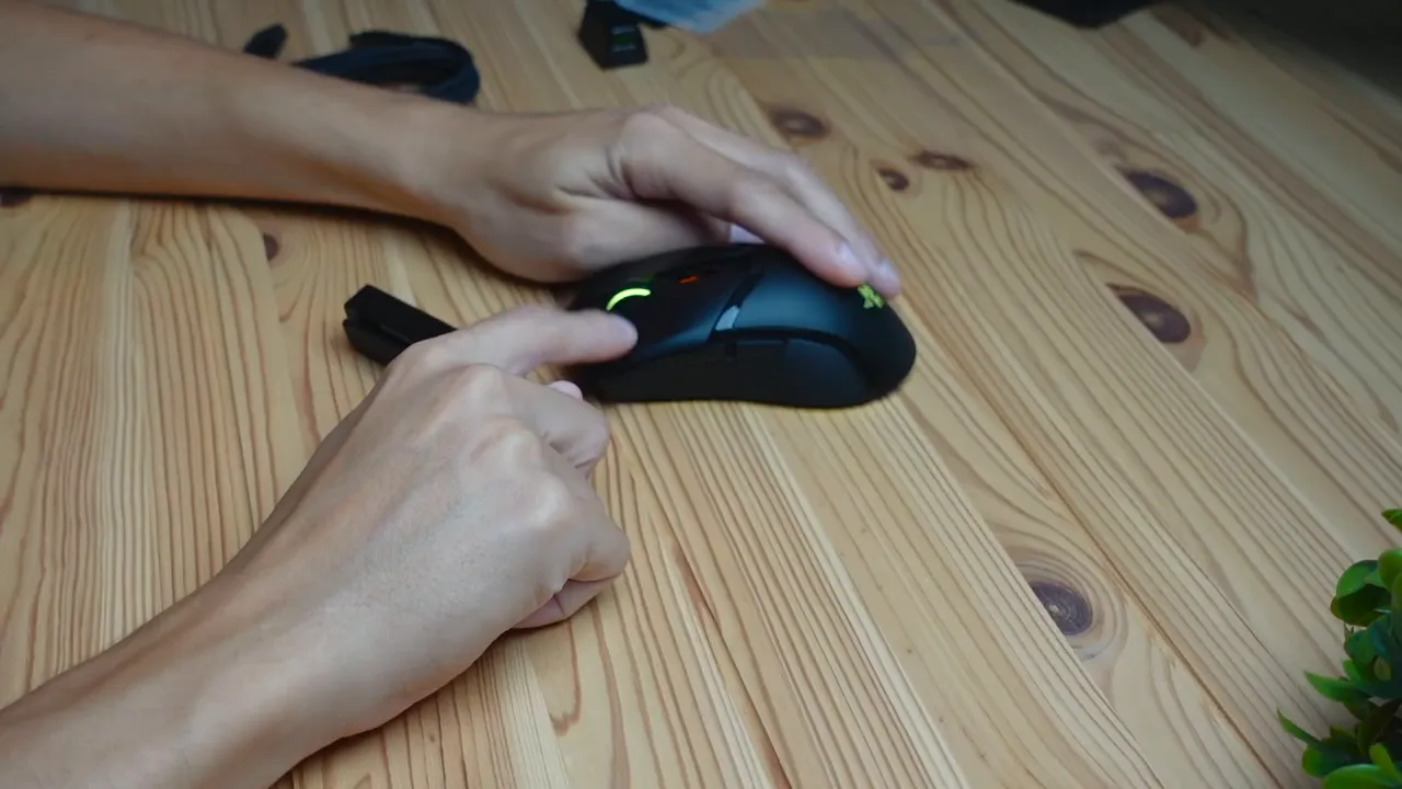 Close-up of a finger pressing the left button of a Razer Cobra Pro mouse showing green RGB lighting and side buttons on a wooden desk