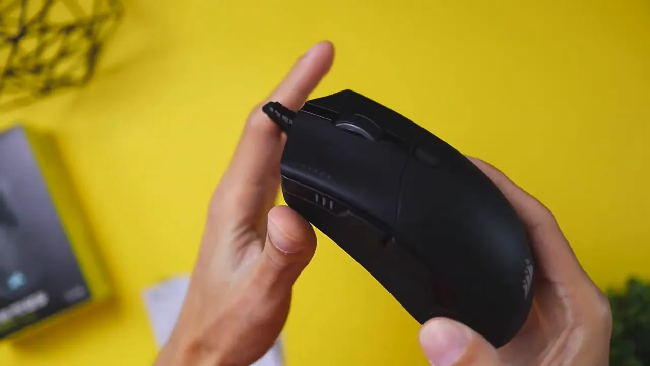 Corsair Sabre RGB Pro gaming mouse held in hands on a yellow background showing side buttons and scroll wheel