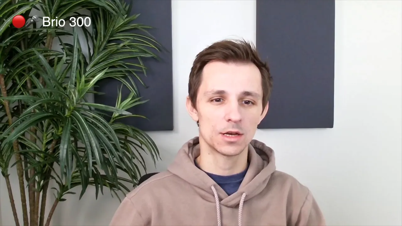 Clear Logitech Brio 300 webcam sample showing a person centered with a plant to the left and acoustic panels behind.