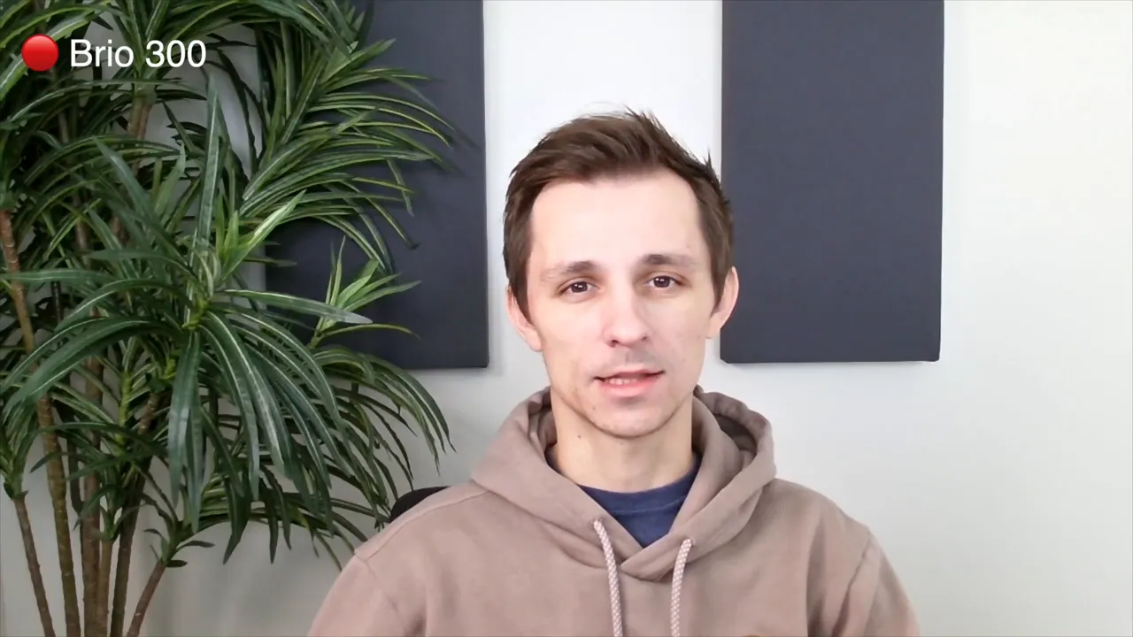 Logitech Brio 300 webcam sample showing the presenter centered with a plant and acoustic panels in the background