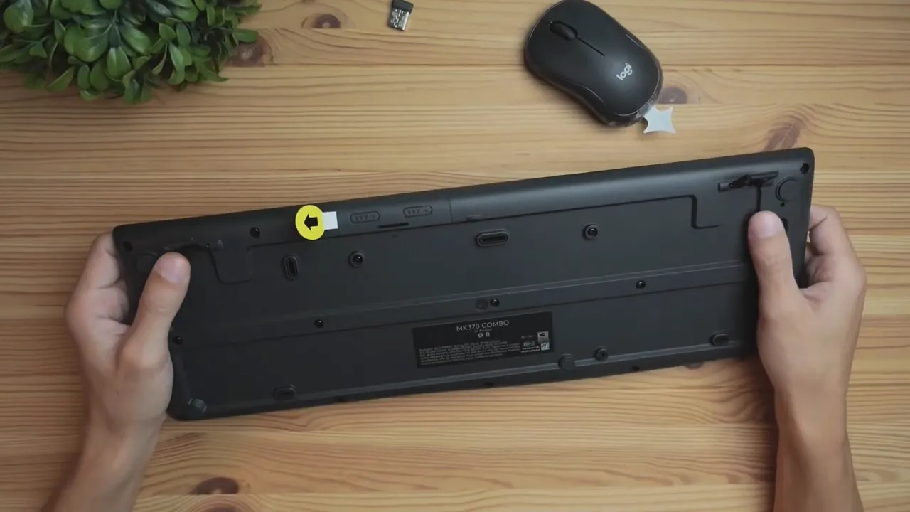 Top-down view of the underside of the Logitech MK370 showing adjustable tilt legs, the receiver slot and the compact mouse on a wooden desk