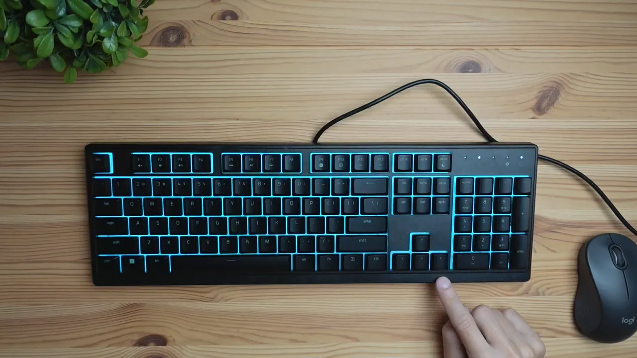Top-down Razer Ornata V3 X keyboard with cyan backlighting on a wooden desk and a finger pressing a key