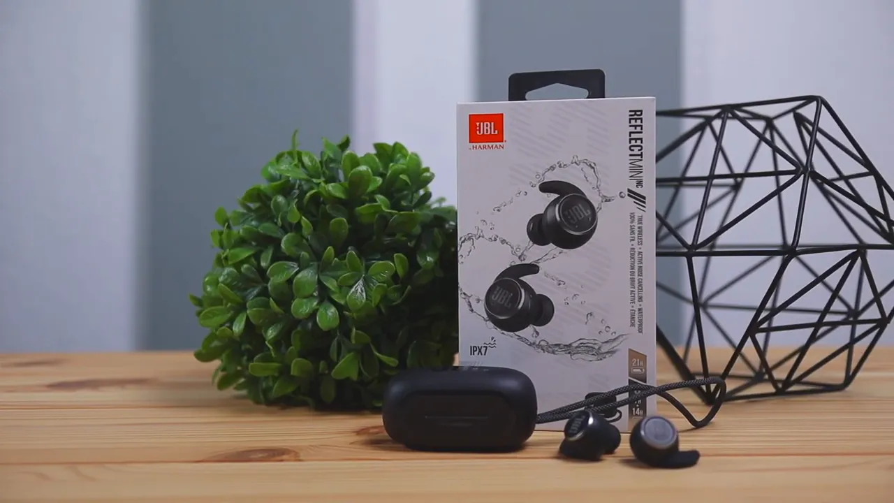 Clear product image of JBL Reflect Mini NC earbuds and charging case with retail box on a wooden table, showing the earbuds' touch surfaces