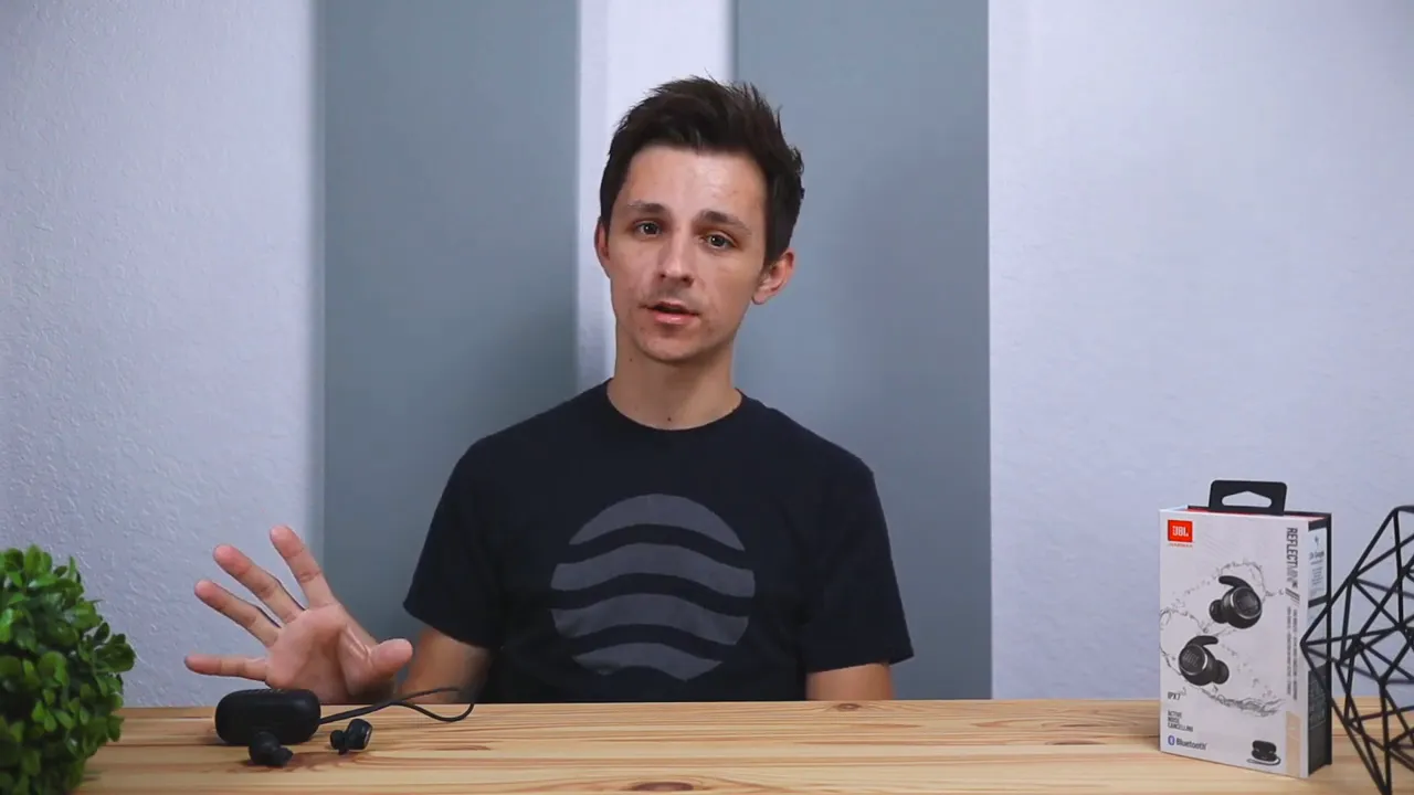 presenter gesturing next to JBL Reflect Mini NC earbuds and packaging on a wooden table
