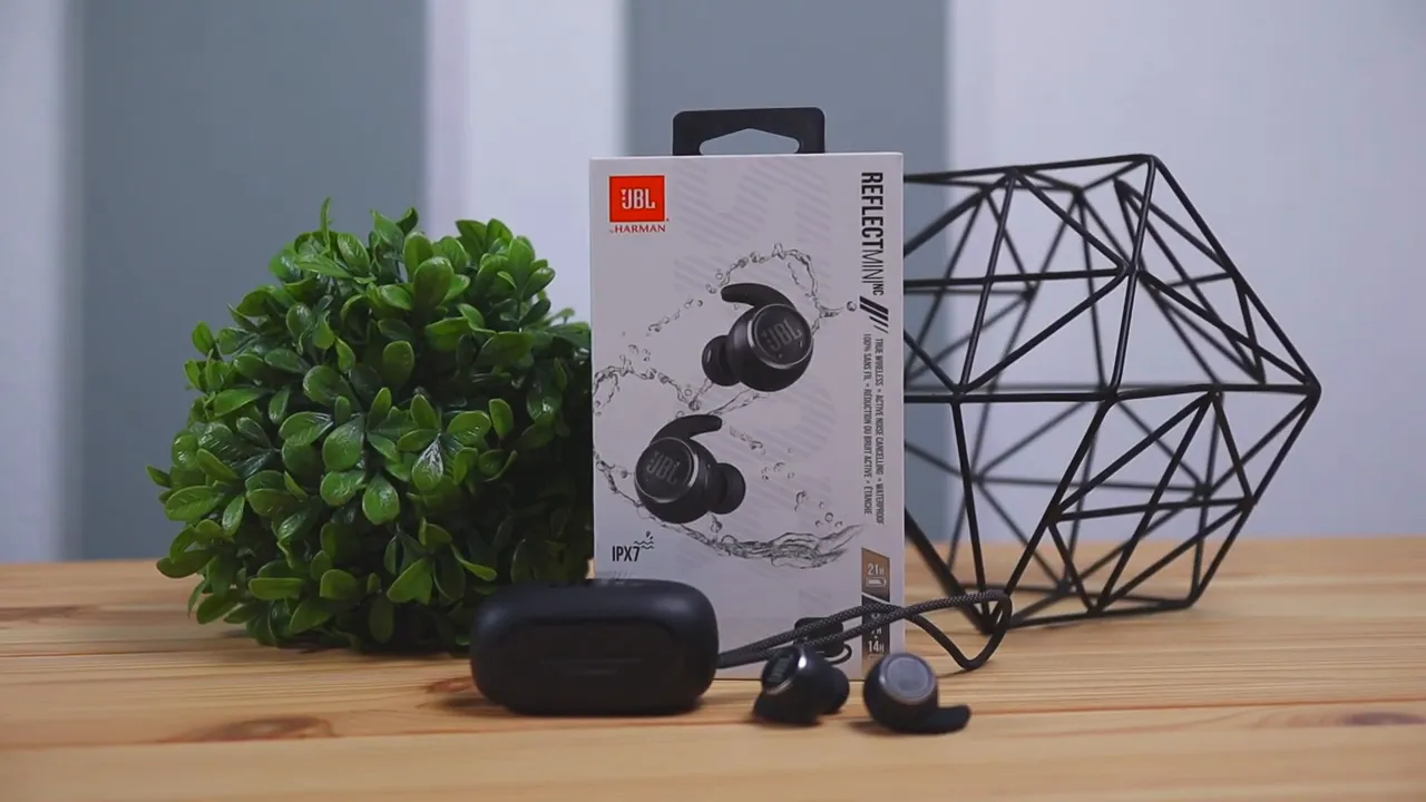 JBL Reflect Mini NC product box with charging case and earbuds on a wooden table with decorative props