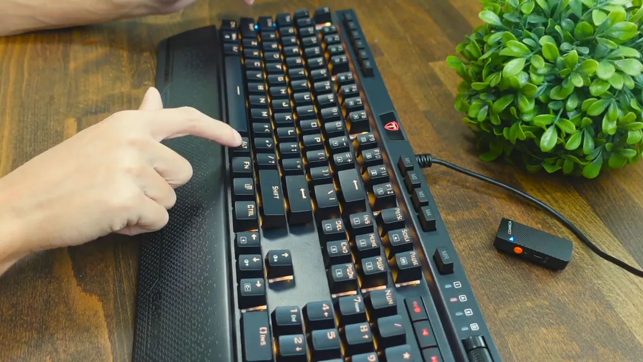 Angled top-down shot of the Risophy full‑size keyboard on a wooden desk with the textured magnetic wrist rest attached; a hand points at the keys, with the USB cable and a small dongle visible.