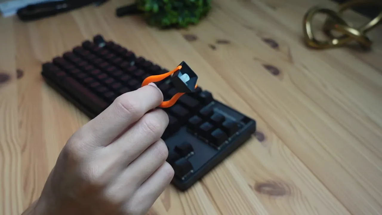 Close-up of an orange keycap puller holding a removed keycap with the Apex 9 TKL keyboard blurred in the background