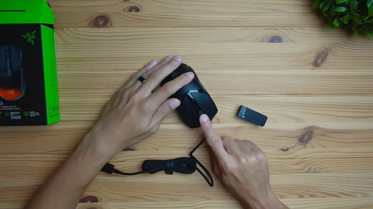 Top-down view of hands pressing the primary button on the Razer Cobra mouse on a wooden desk with the box nearby.