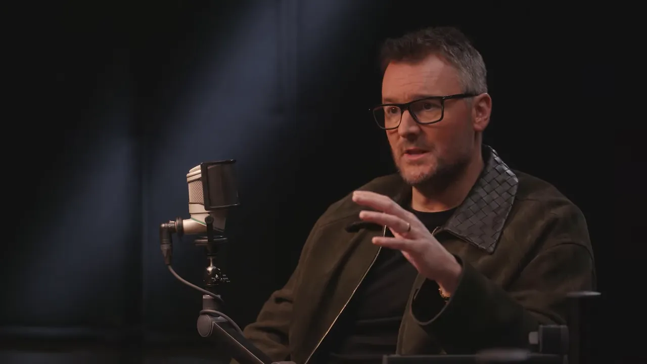 Interview subject in a studio interview, speaking into a microphone with a hand gesture, clear lighting and focus.