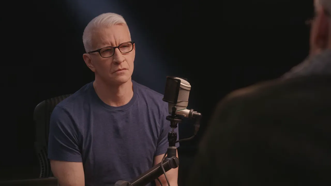 studio interviewer listening across from a guest with a microphone between them, serious expression
