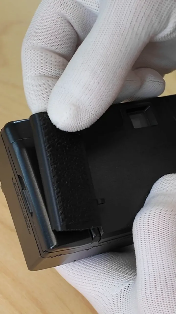 Gros plan sur la charnière du compartiment batterie du Yashica T3 avec le verrou en plastique cassé et le couvercle de remplacement, tenu par un gant blanc.