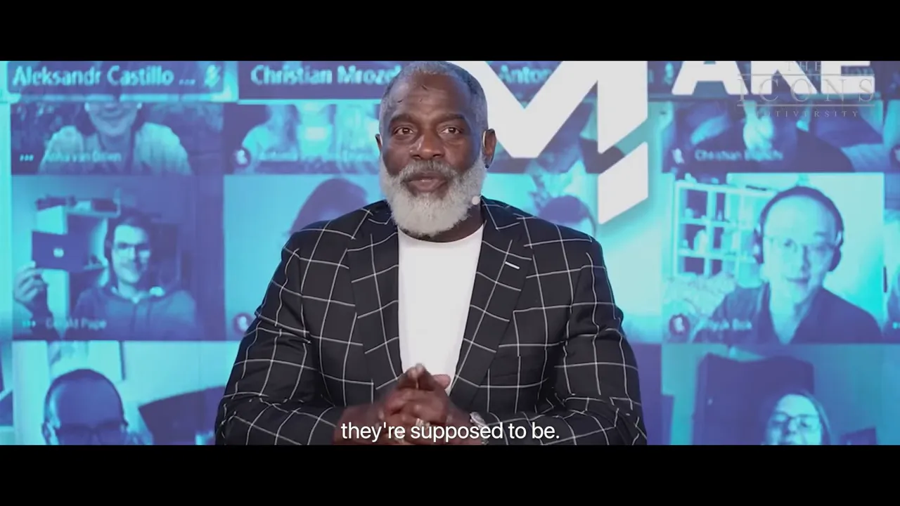 Presenter in a checkered blazer clasping hands and speaking in front of a blue virtual audience wall with subtitle 'they're supposed to be'.