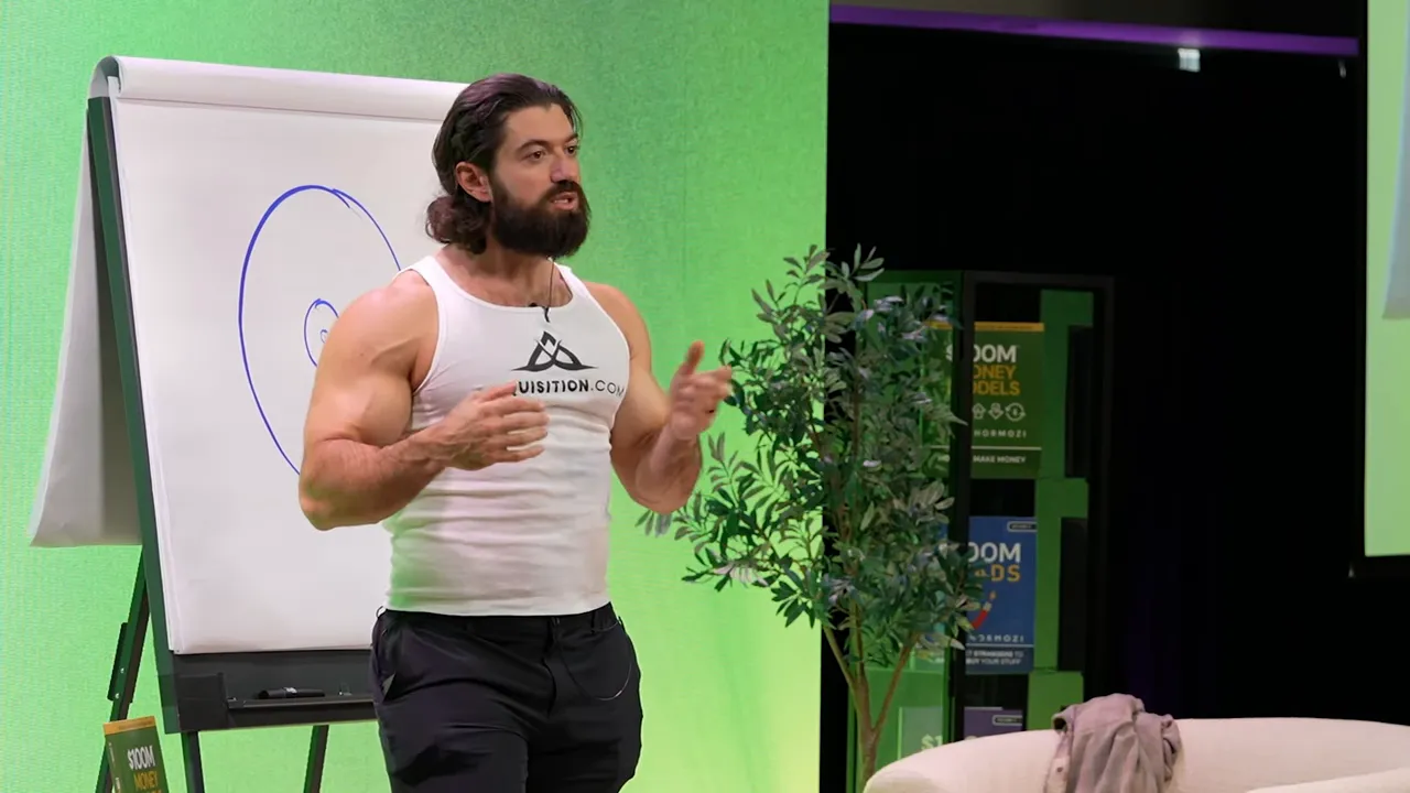 Talk-stage shot showing a presenter beside a flipchart with a target drawn, speaking and using hand gestures to emphasize a point.