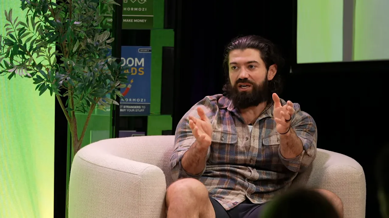 Presenter seated onstage in an armchair, leaning forward and gesturing with both hands as if describing a sequence or steps.