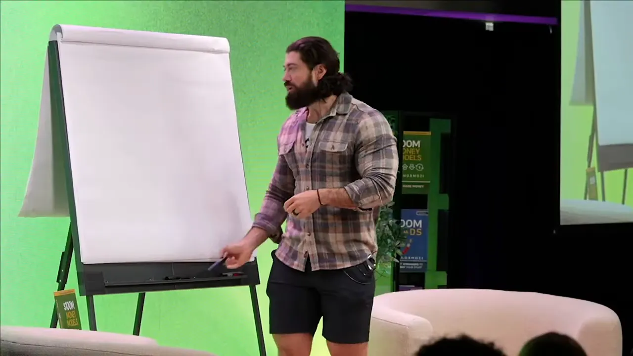 Presenter standing beside a large blank flipchart on stage, holding a marker and pointing toward the board while speaking to the audience.