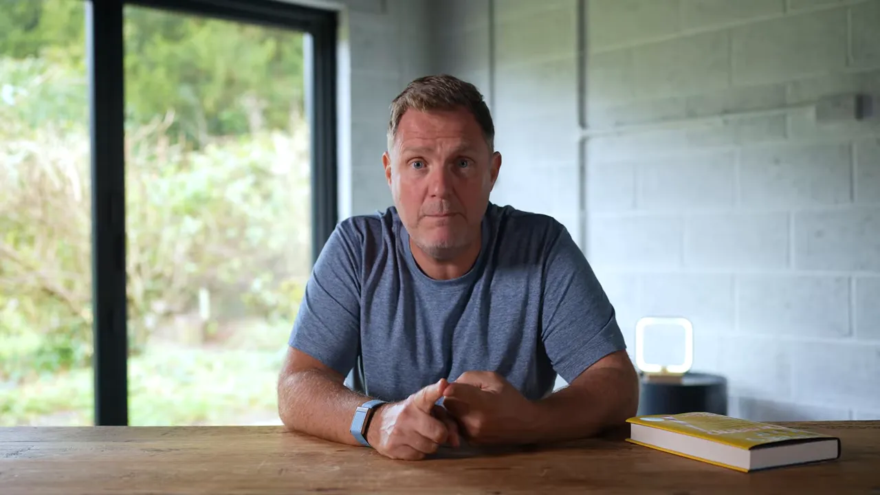 Centered medium shot of presenter at a wooden table with a book to the side, speaking directly to camera