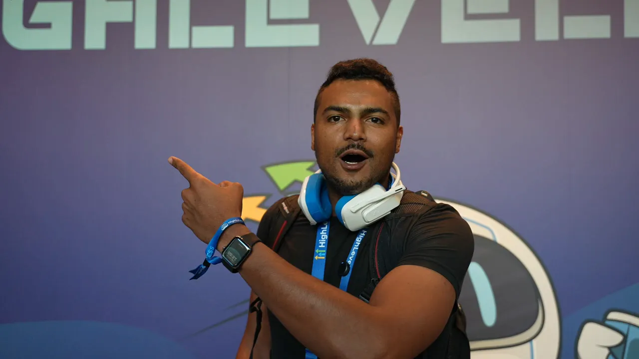 Conference attendee with blue-and-white headphones and lanyard pointing and speaking in front of a purple-branded background