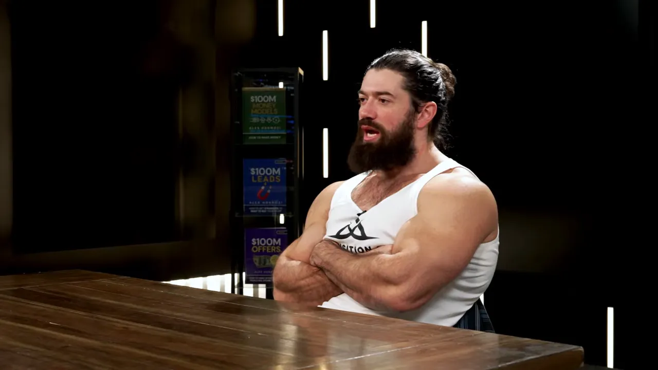 Side-angle of presenter mid-sentence with arms crossed at a wooden table, studio lights in background — clear and well-composed