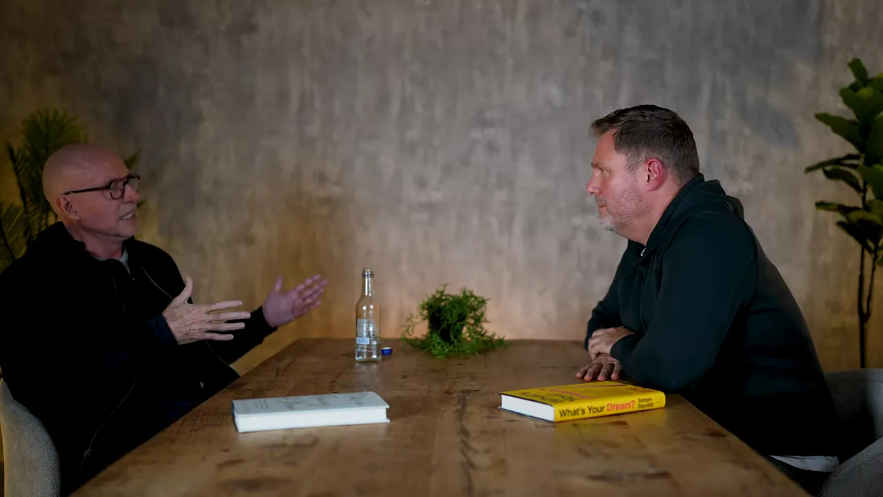 Wide shot of two people seated across a wooden table in discussion, books and bottle on the table