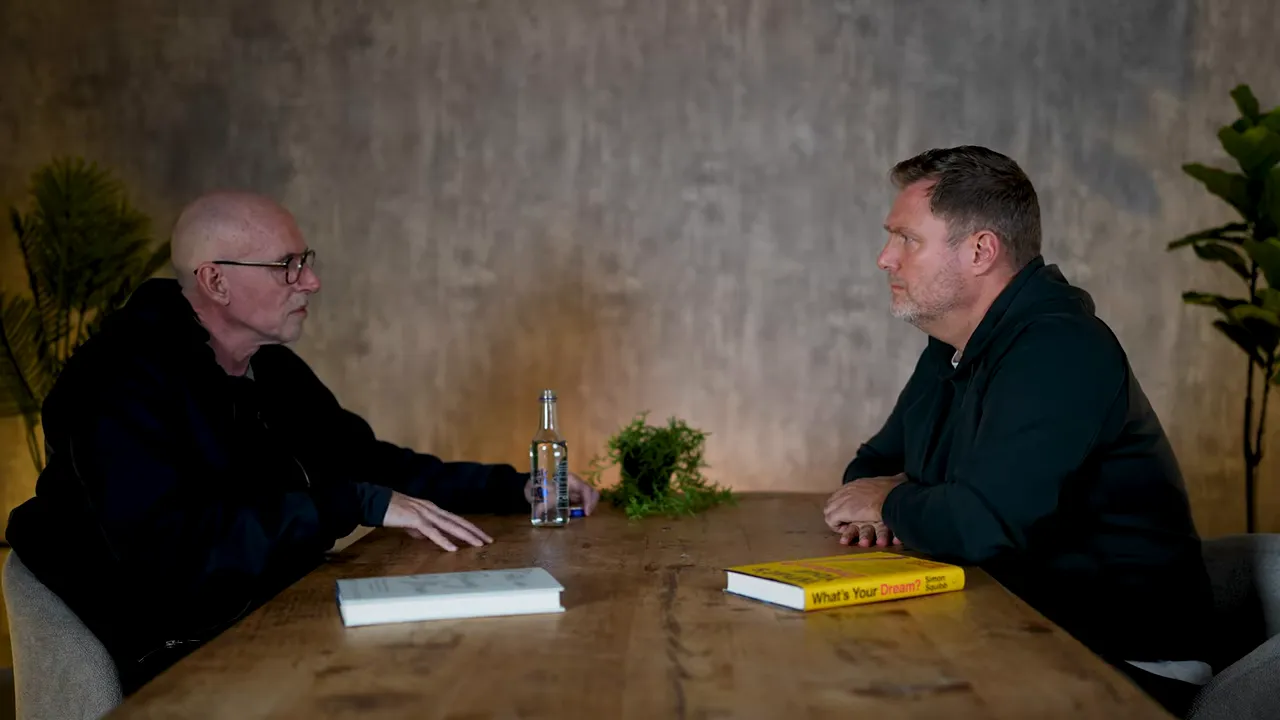 Wide balanced shot of two people facing each other across a wooden table in conversation with a book and plant visible