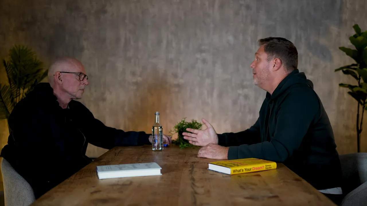 Wide, well‑lit two-person interview shot across a wooden table with a book and water bottle; one person gesturing while explaining
