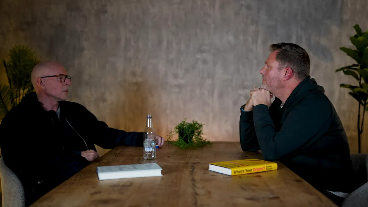 Wide view of two people seated across a wooden table in conversation with a book and a water bottle between them and plants in the background