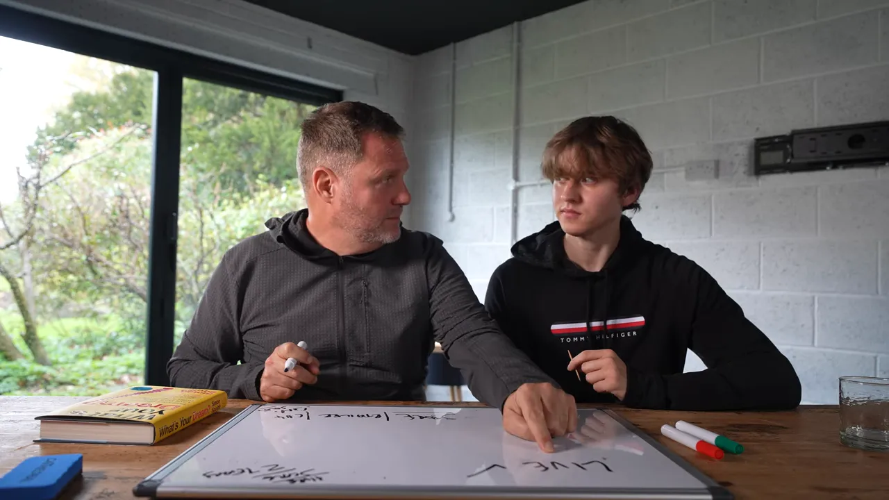 Two people at a table pointing to and discussing notes on a whiteboard with markers and a book nearby.