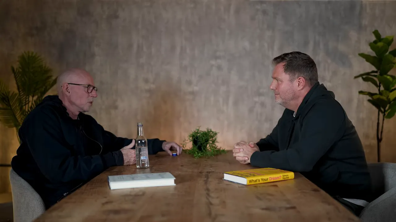 Balanced wide shot of two people facing each other across a wooden table, with a book and plant visible — a scene suggesting mentorship and focused conversation.