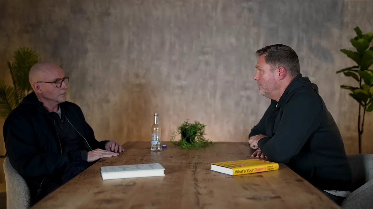 Wide, high-quality shot of two speakers seated across a wooden table facing each other, with books, a bottle, and a plant — a concluding discussion.