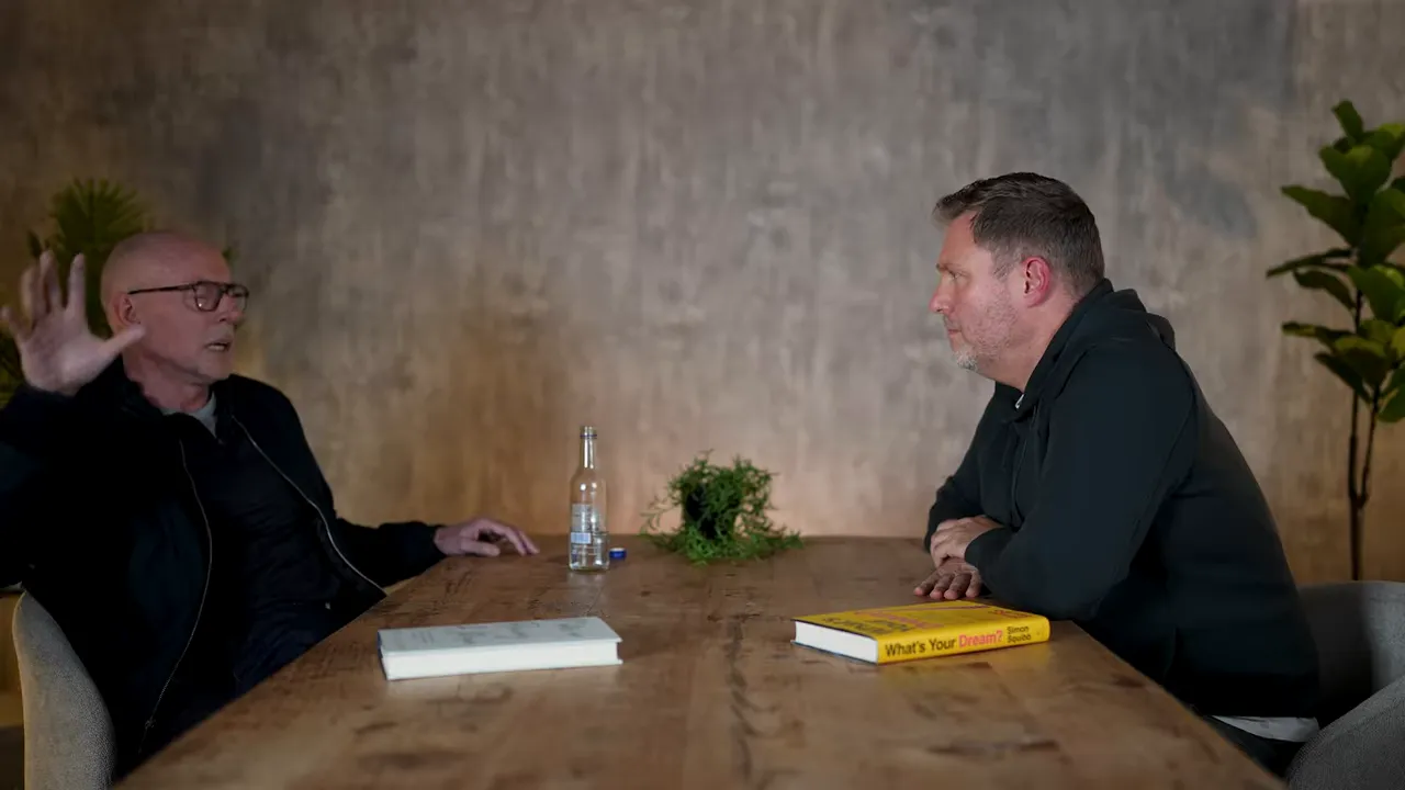 High-quality wide shot of two speakers at a wooden table facing each other, with a book, bottle, and plant on the table.