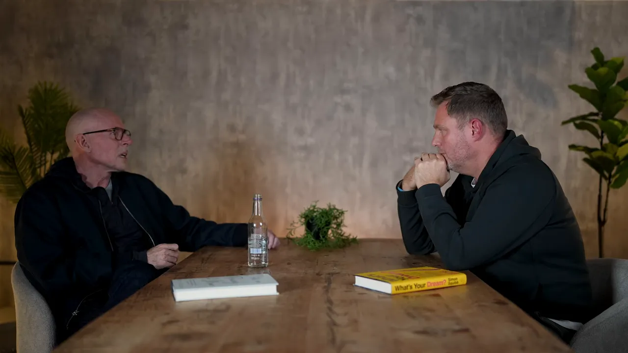 High-quality wide shot of two speakers across a wooden table, a book and water bottle on the table and a neutral textured wall behind them.
