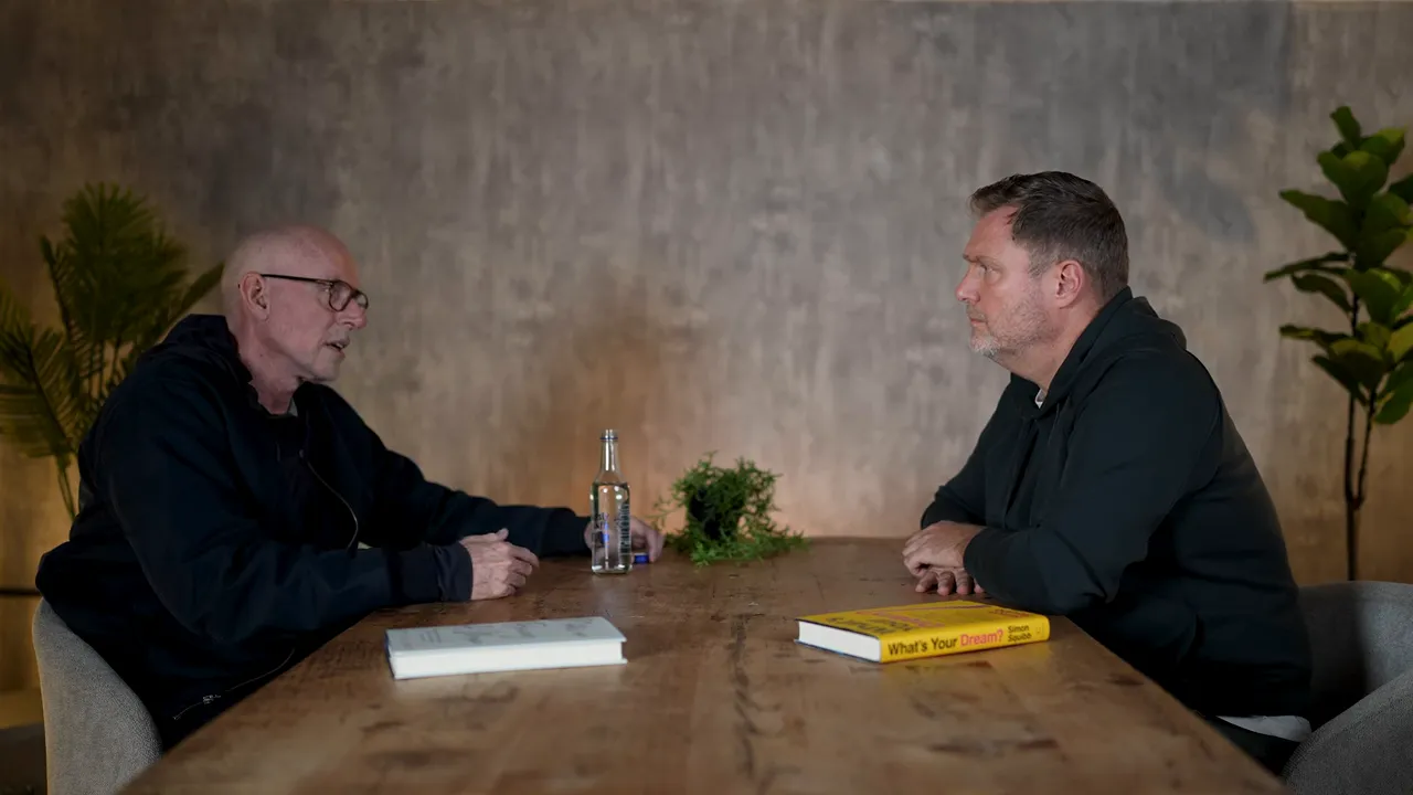 Wide, high-quality shot of two people seated across a wooden table, facing each other with books and a water bottle on the table.