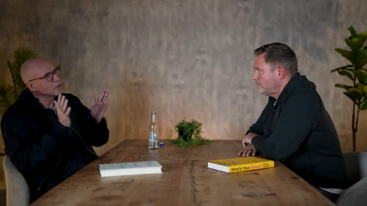 Wide interview shot showing two speakers across a wooden table, one gesturing while the other listens, with a book and plant visible.