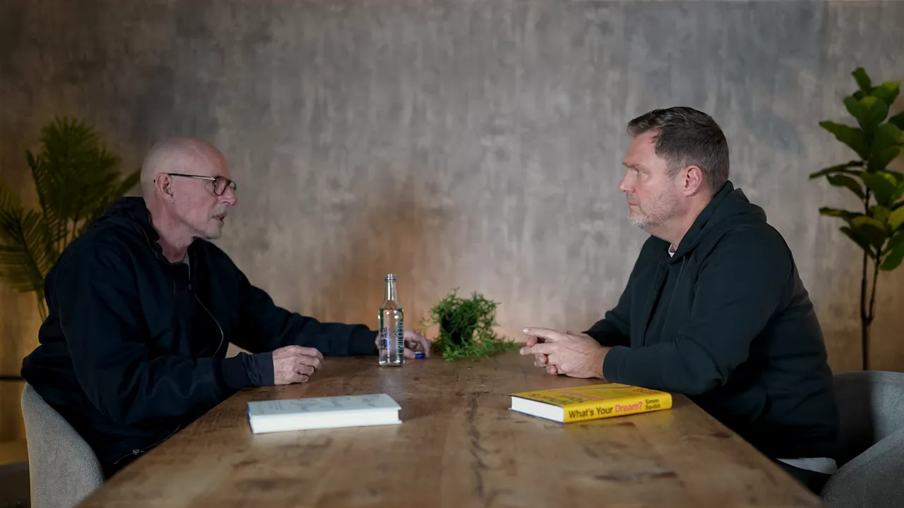 Two speakers in a business interview across a wooden table with a visible book and water bottle, illustrating a discussion about systems and tools.