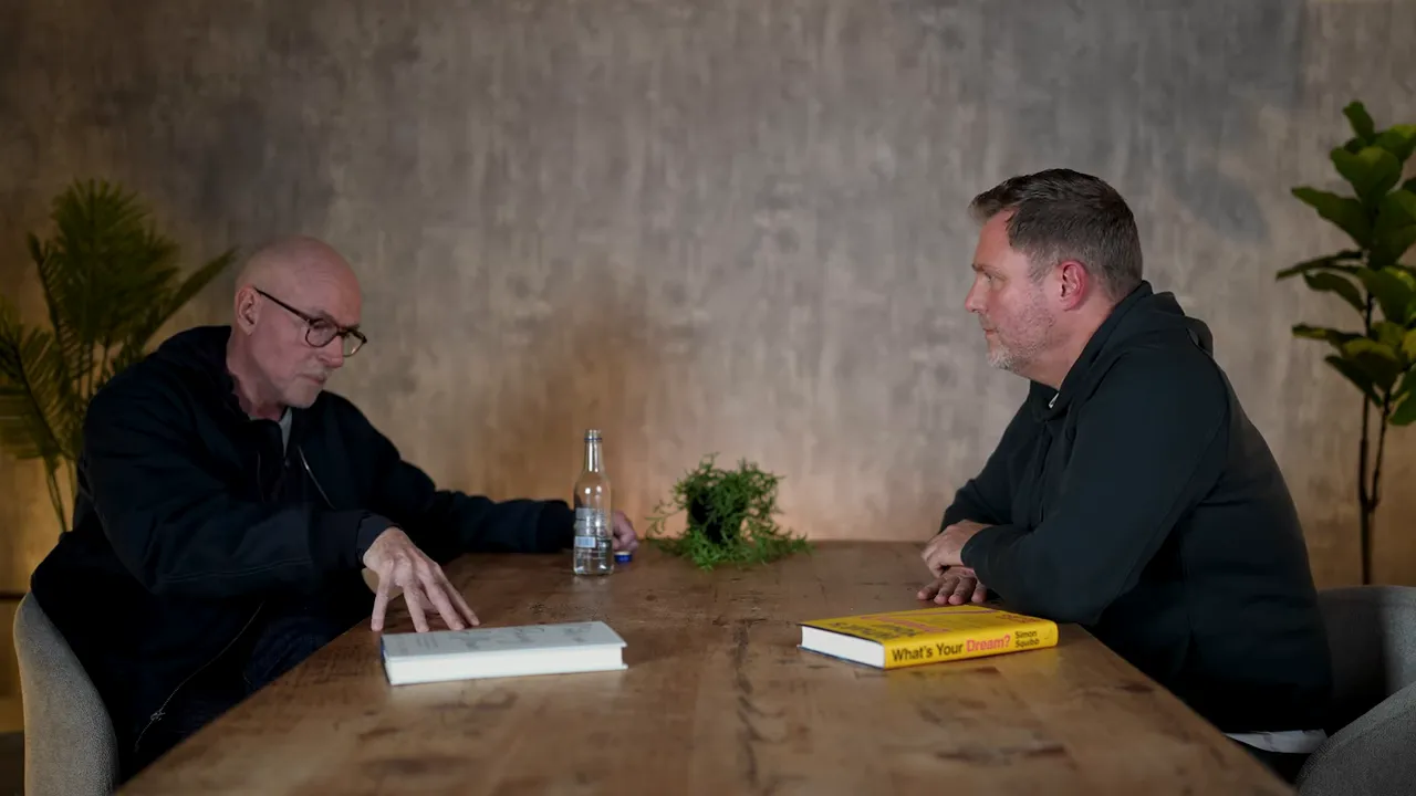 wide interview shot with one person gesturing and a yellow book on the table
