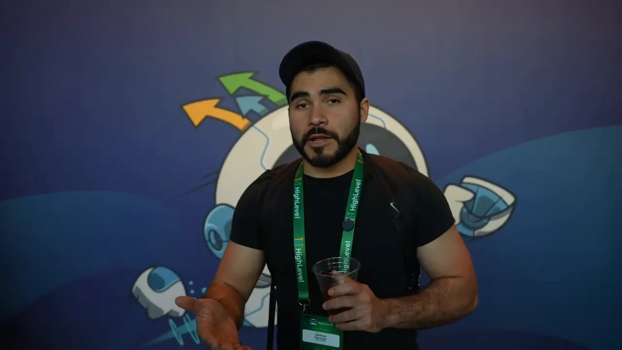 conference speaker gesturing while holding a drink, wearing a green lanyard, with a blue illustrated backdrop