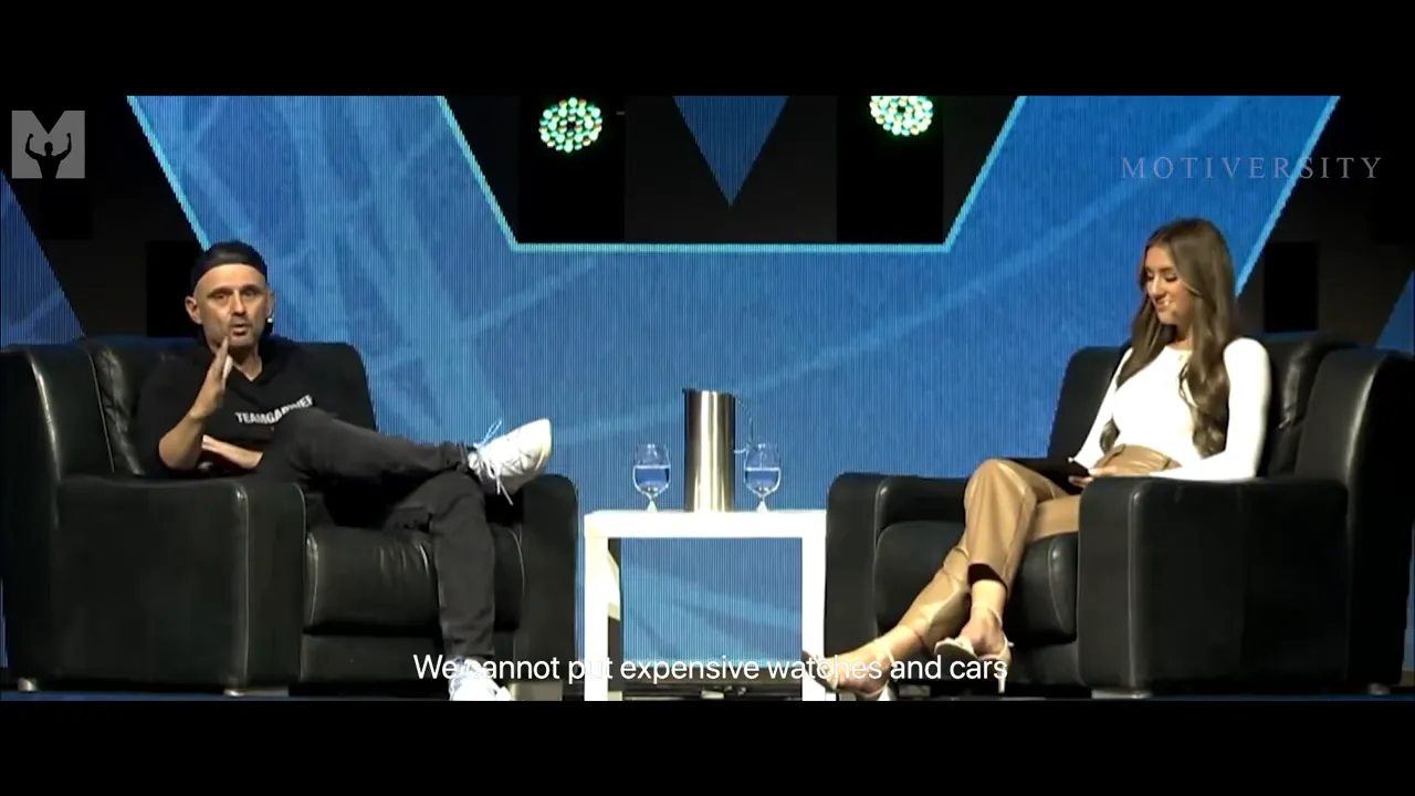 Speaker gesturing while seated on stage during an interview with a blue backdrop and interviewer on the right