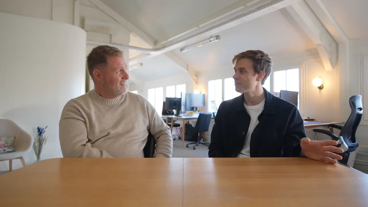 Two colleagues at a table in an office, one gesturing with his hand while they talk